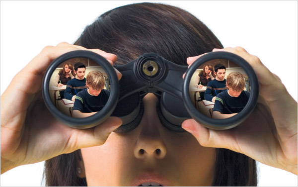 A photograph of a close-up of a woman holding binoculars up to her eyes.  The reflection in the lenses shows students sitting in a classroom.