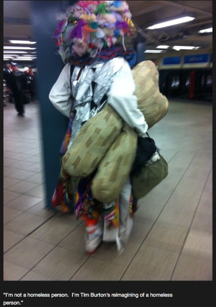 A photograph of a person in clownish garb holding a stuffed toy that is vaguely shaped like a human chromosome. He/she is wearing a giant bulbous wig made of colored pieces of fabric. The caption provided says "I'm not a homeless person. I'm Tim Burton's reimagining of a homeless person."