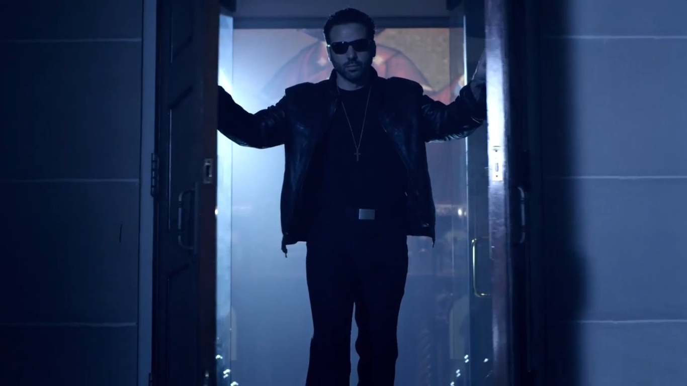 Image of Jamie Casino opening double wooden doors to a church, standing between them, while wearing a leather jacket and sunglasses.