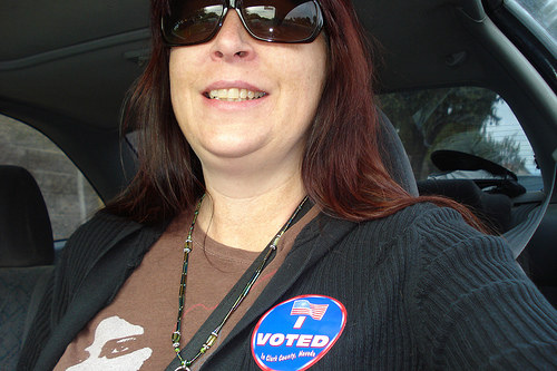 Woman wears large circular "I Voted" sticker on shirt while sitting in a car