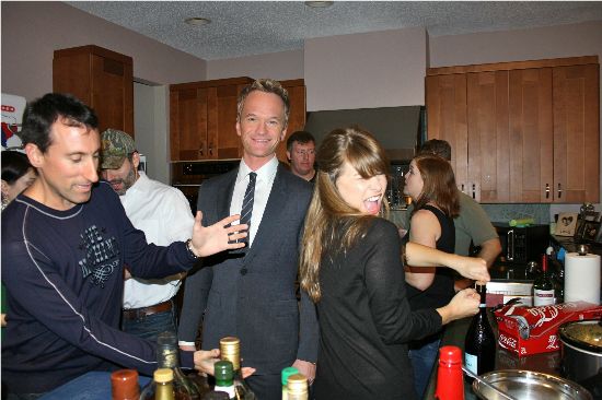 This image depicts Neal Patrick Harris in a suit posed between two drunk people; on the right foreground stands a girl in a black dress posing with her back to the camera looking over her shoulder; to the left foreground a man gestures towards her backside.