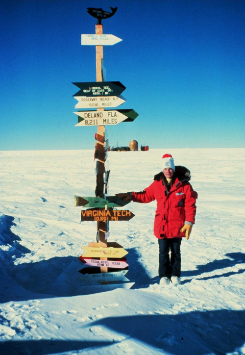 Antarctic researcher stands at sign post with signs stating distance to different locations