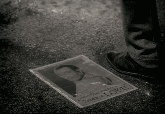 The image is of a headshot of George Valentin in a white suit, dressed as his character from his film Tears of Love.  The headshot lies on the wet ground as a foot stands near it.